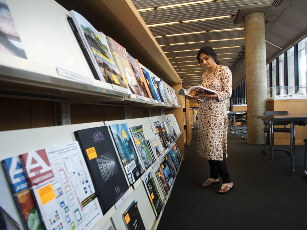 in loeb library a student is looking at a book that she is holding in her hands  standing in front of a shelf of books