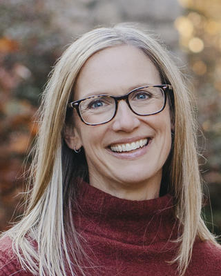 Nicole Breen faces the camera with a warm, open smile, wearing rectangular tortoiseshell glasses and a burgundy turtleneck sweater. Her portrait is set against a softly blurred, natural outdoor background featuring autumnal foliage